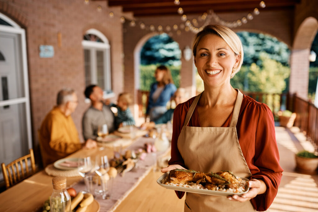 Happy woman serving food to her extended family on a patio and looking at camera.